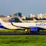 A side profile of an IndiGo Airbus A320-271N, registration VT-IZI, taxiing or beginning its takeoff roll on a runway. The aircraft's white and blue livery is bright under the clear sky, and the background features airport structures and several parked Biman Bangladesh Airlines aircraft with their red and white tail logos. Green grass lines the foreground of the runway.