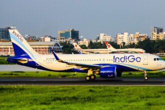 A side profile of an IndiGo Airbus A320-271N, registration VT-IZI, taxiing or beginning its takeoff roll on a runway. The aircraft's white and blue livery is bright under the clear sky, and the background features airport structures and several parked Biman Bangladesh Airlines aircraft with their red and white tail logos. Green grass lines the foreground of the runway.