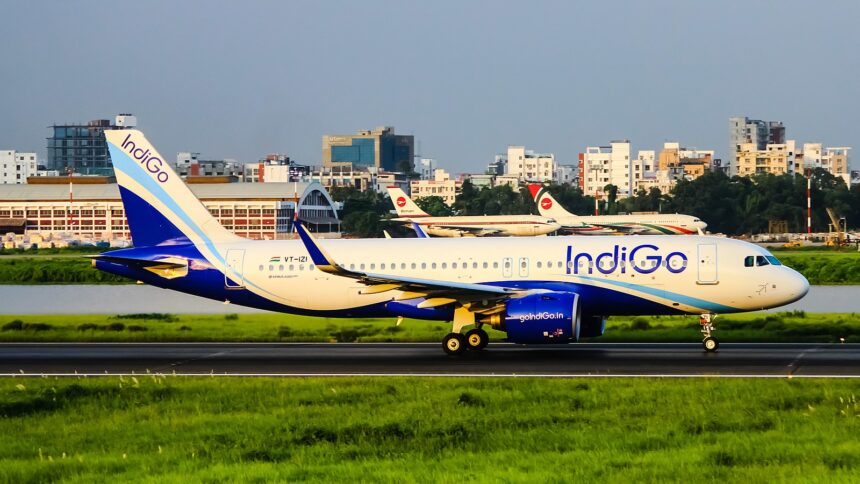 A side profile of an IndiGo Airbus A320-271N, registration VT-IZI, taxiing or beginning its takeoff roll on a runway. The aircraft's white and blue livery is bright under the clear sky, and the background features airport structures and several parked Biman Bangladesh Airlines aircraft with their red and white tail logos. Green grass lines the foreground of the runway.