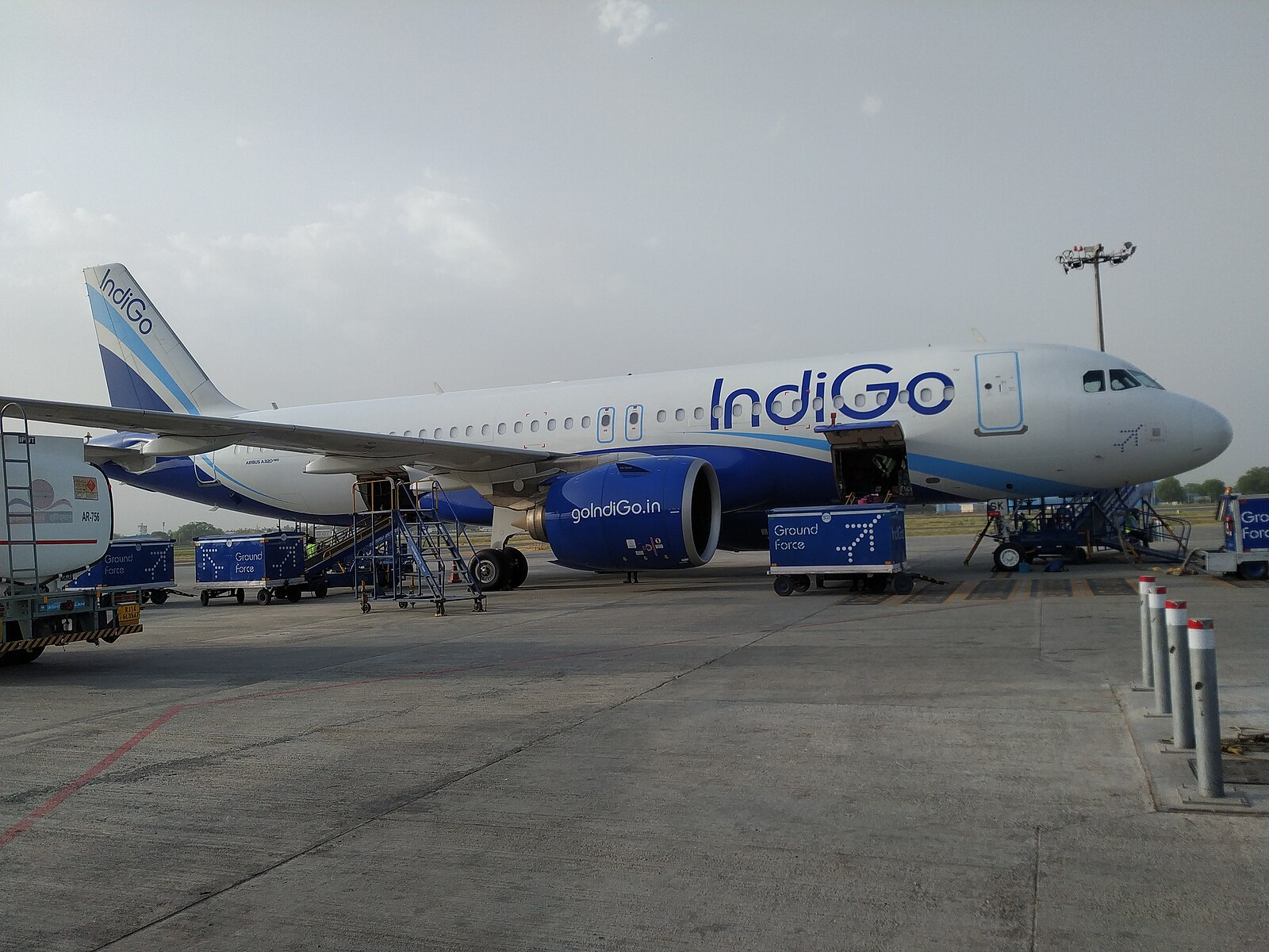 An IndiGo Airbus A320 aircraft parked on the tarmac at Delhi Airport. The plane is in the airline’s signature white and blue livery, with ground support equipment and luggage carts nearby. Terminal structures and other parked aircraft are visible in the background.