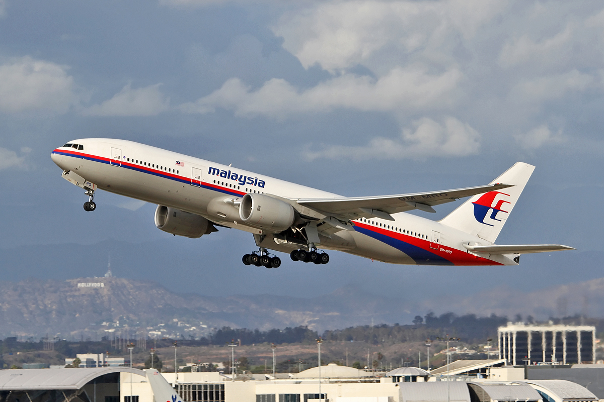 A Malaysia Airlines Boeing 777-200ER (registration 9M-MRO) taking off from an airport. The aircraft is white with red and blue stripes along the fuselage and the airline's red and blue logo on the tail. In the background, a hilly landscape features the iconic Hollywood sign, and the landing gear is in the process of retracting against a cloudy sky.