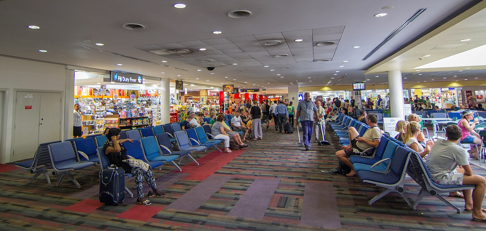 A wide view of a crowded airport terminal featuring rows of blue tandem seating occupied by travelers. The floor is covered in a patterned carpet with red and purple accents. To the left, a 