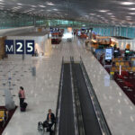 An interior view looking down a long, modern airport concourse at Paris Charles de Gaulle Airport. The terminal features a high, vaulted ceiling with a metallic grid pattern and polished white floors. Large signs hanging from the ceiling indicate gates "M25," "M26," and "M27." To the right, there are seating areas with rows of orange and red chairs, while a moving walkway runs down the center of the hall. A few passengers and staff members are scattered throughout the spacious terminal.