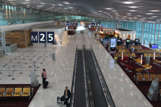 An interior view looking down a long, modern airport concourse at Paris Charles de Gaulle Airport. The terminal features a high, vaulted ceiling with a metallic grid pattern and polished white floors. Large signs hanging from the ceiling indicate gates "M25," "M26," and "M27." To the right, there are seating areas with rows of orange and red chairs, while a moving walkway runs down the center of the hall. A few passengers and staff members are scattered throughout the spacious terminal.