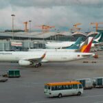 A Philippine Airlines Airbus A321 is parked at an airport gate at Hong Kong International Airport under an overcast sky. In the background, other aircraft from Cathay Pacific and XiamenAir are visible alongside the terminal's distinctive wavy silver roof. The foreground shows the tarmac with ground service equipment, a white and yellow shuttle bus, and cargo containers, while several construction cranes rise in the distance.