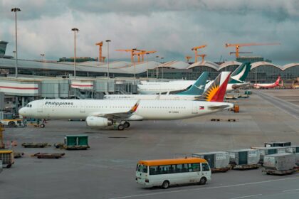 A Philippine Airlines Airbus A321 is parked at an airport gate at Hong Kong International Airport under an overcast sky. In the background, other aircraft from Cathay Pacific and XiamenAir are visible alongside the terminal's distinctive wavy silver roof. The foreground shows the tarmac with ground service equipment, a white and yellow shuttle bus, and cargo containers, while several construction cranes rise in the distance.