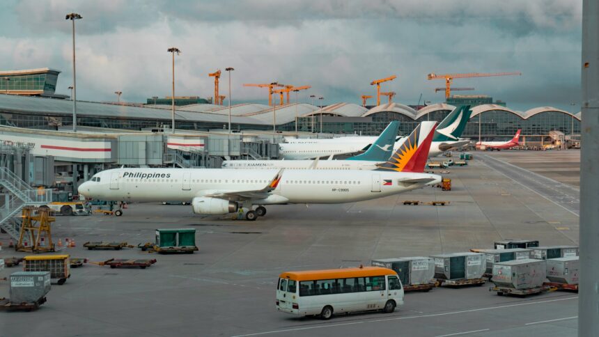 A Philippine Airlines Airbus A321 is parked at an airport gate at Hong Kong International Airport under an overcast sky. In the background, other aircraft from Cathay Pacific and XiamenAir are visible alongside the terminal's distinctive wavy silver roof. The foreground shows the tarmac with ground service equipment, a white and yellow shuttle bus, and cargo containers, while several construction cranes rise in the distance.