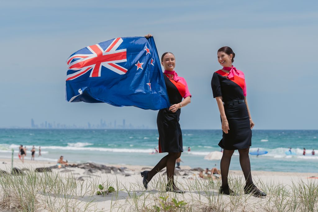 Two women in Qantas cabin crew uniforms hold a New Zealand flag at a beach.