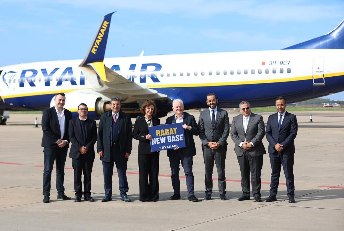 Eight individuals in suits (seven men and one woman) stand in front of a Ryanair jetliner, with the man and woman in the middle holding a card that reads "RABAT NEW BASE RYANAIR".
