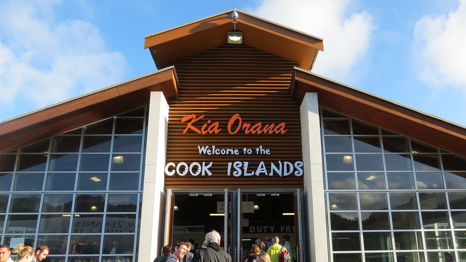 The exterior of Rarotonga International Airport terminal under a blue sky with scattered clouds. The building features large glass windows and a prominent wooden A-frame central section. Orange and white lettering on the facade reads 