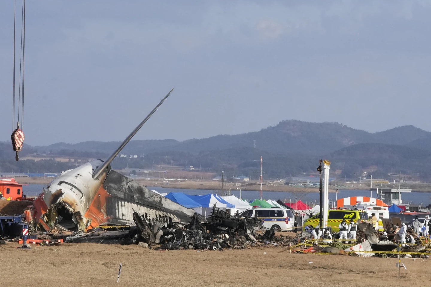 A photograph from Monday, December 30, 2024, showing the charred and mangled wreckage of Jeju Air Flight 2216 at Muan International Airport. Several emergency responders in yellow protective suits and red helmets are conducting search and salvage operations among the debris. The aircraft's orange and white tail section is tilted sharply upward, supported by thick orange lifting straps. The fuselage is heavily damaged, showing exposed metal and significant fire blackening. In the background, a red fire truck and other emergency vehicles are stationed behind a perimeter of barbed-wire fencing under a grey, overcast sky.