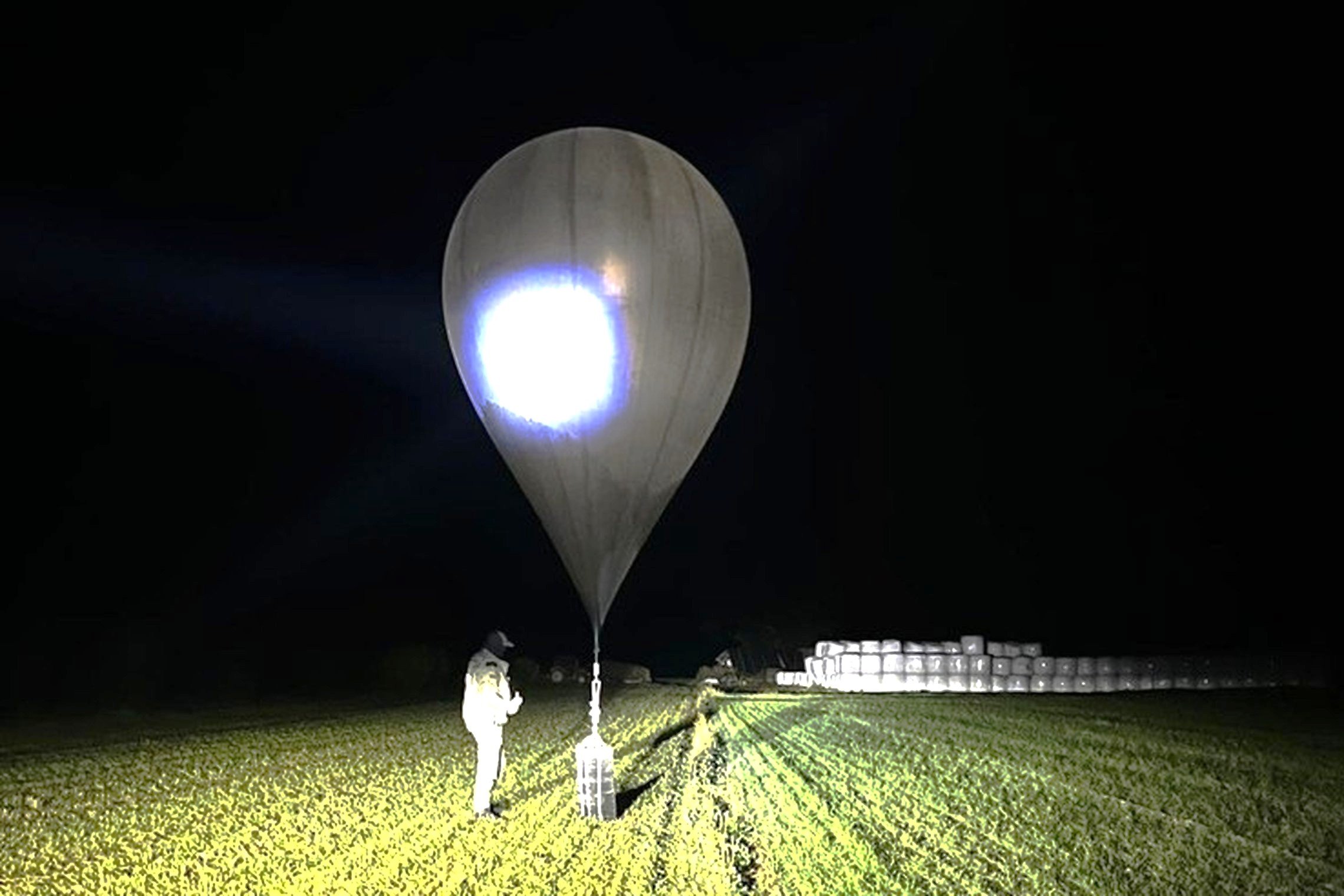 A border guard approaches a weather balloon on a grass field, seemingly carrying contraband, at night. 
