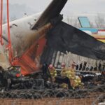 A photograph of emergency responders in yellow uniforms and red helmets inspecting the charred, mangled wreckage of a Jeju Air plane. The tail section is tilted upward, held by orange crane straps, while the fuselage shows significant fire damage and exposed metal. Fire trucks and emergency vehicles are visible in the background behind a perimeter of barbed wire.