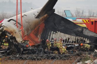 A photograph of emergency responders in yellow uniforms and red helmets inspecting the charred, mangled wreckage of a Jeju Air plane. The tail section is tilted upward, held by orange crane straps, while the fuselage shows significant fire damage and exposed metal. Fire trucks and emergency vehicles are visible in the background behind a perimeter of barbed wire.