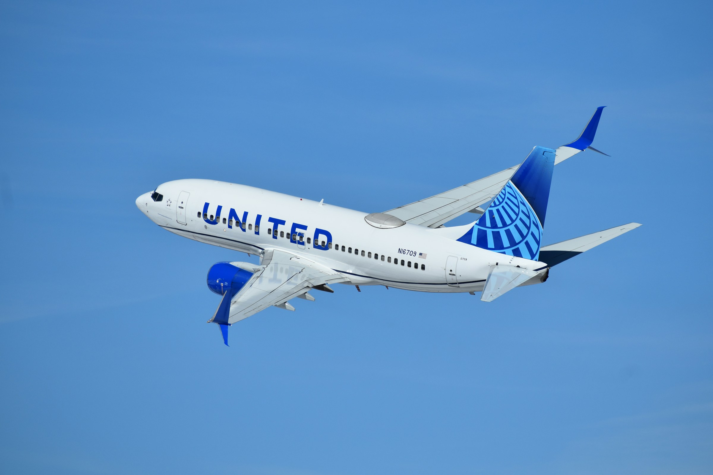 A United Airlines Boeing 737 aircraft, tail number N16709, is shown climbing into a clear blue sky. The plane is viewed from a slightly rear-underneath angle, showcasing the blue United livery on the engines, winglets, and the globe logo on the tail fin. The landing gear is retracted, and the aircraft is banked slightly to the left.