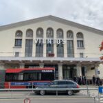 The front of the Vilnius Airport building's entrance with a bus and a car passing through.