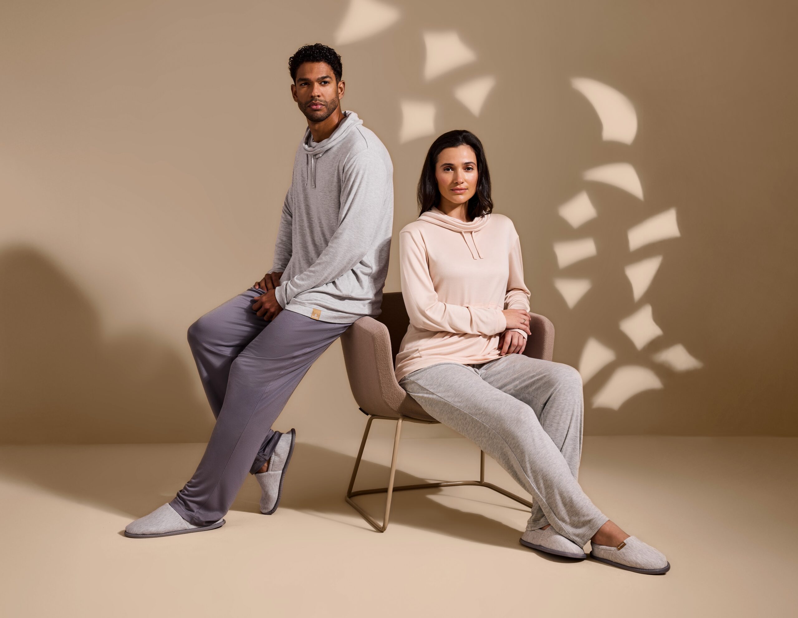 Emirates launches new luxury loungewear. Male modeal leanning against chair. He's wearing a grey hoodie and lounge pants from the new luxury loungeware collection. The female model is seated and wearing a pink hoodie jumper and grey lounge pants.