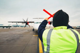 A ground crew worker in a high-visibility jacket guides a turboprop aircraft on the apron at Coventry Airport.