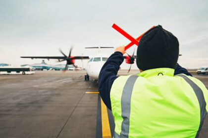 A ground crew worker in a high-visibility jacket guides a turboprop aircraft on the apron at Coventry Airport.