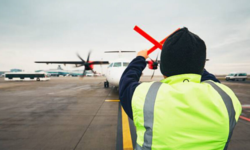 A ground crew worker in a high-visibility jacket guides a turboprop aircraft on the apron at Coventry Airport.