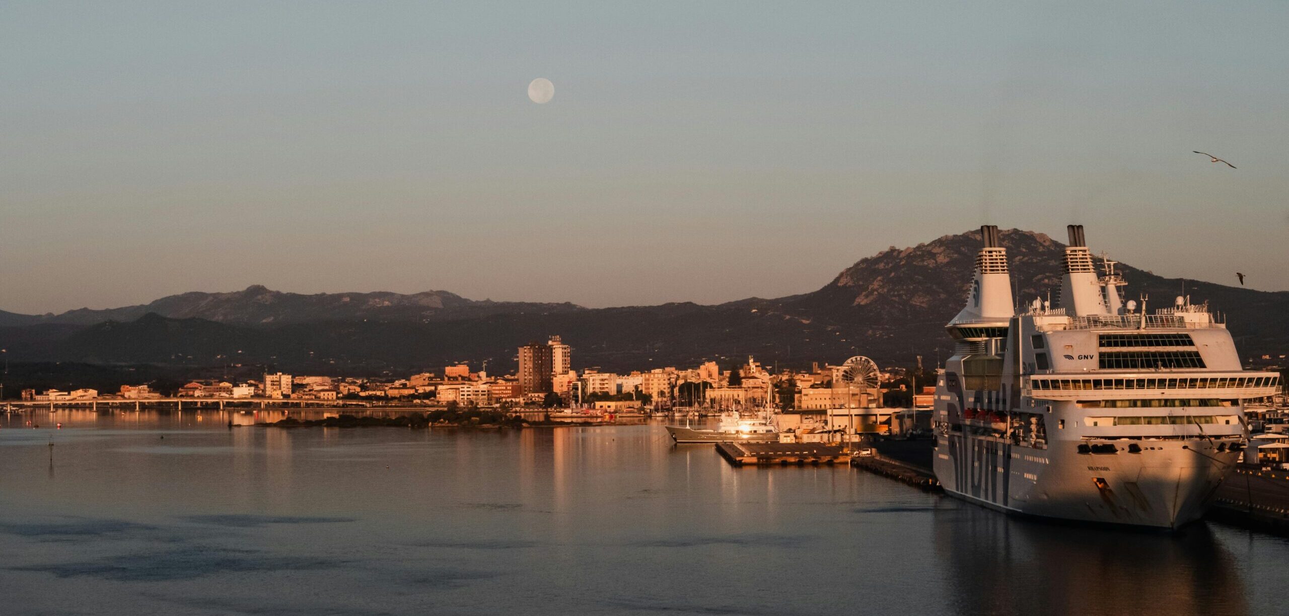 Olbia from the harbour at dusk. City lights and tall white buildings are surrounded by mountains and water. The moon is visible