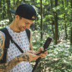 A guy walking through a forest, holding a phone and a camera