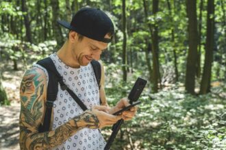 A guy walking through a forest, holding a phone and a camera