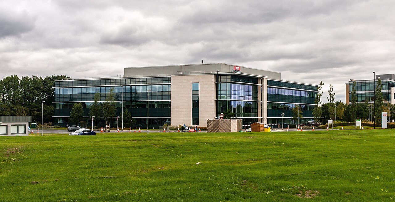 A view of Manchester Airport's main terminal, with an area of grass being in the forefront of the photo and cars being able to be seen just behind this area in front of the airport. 
