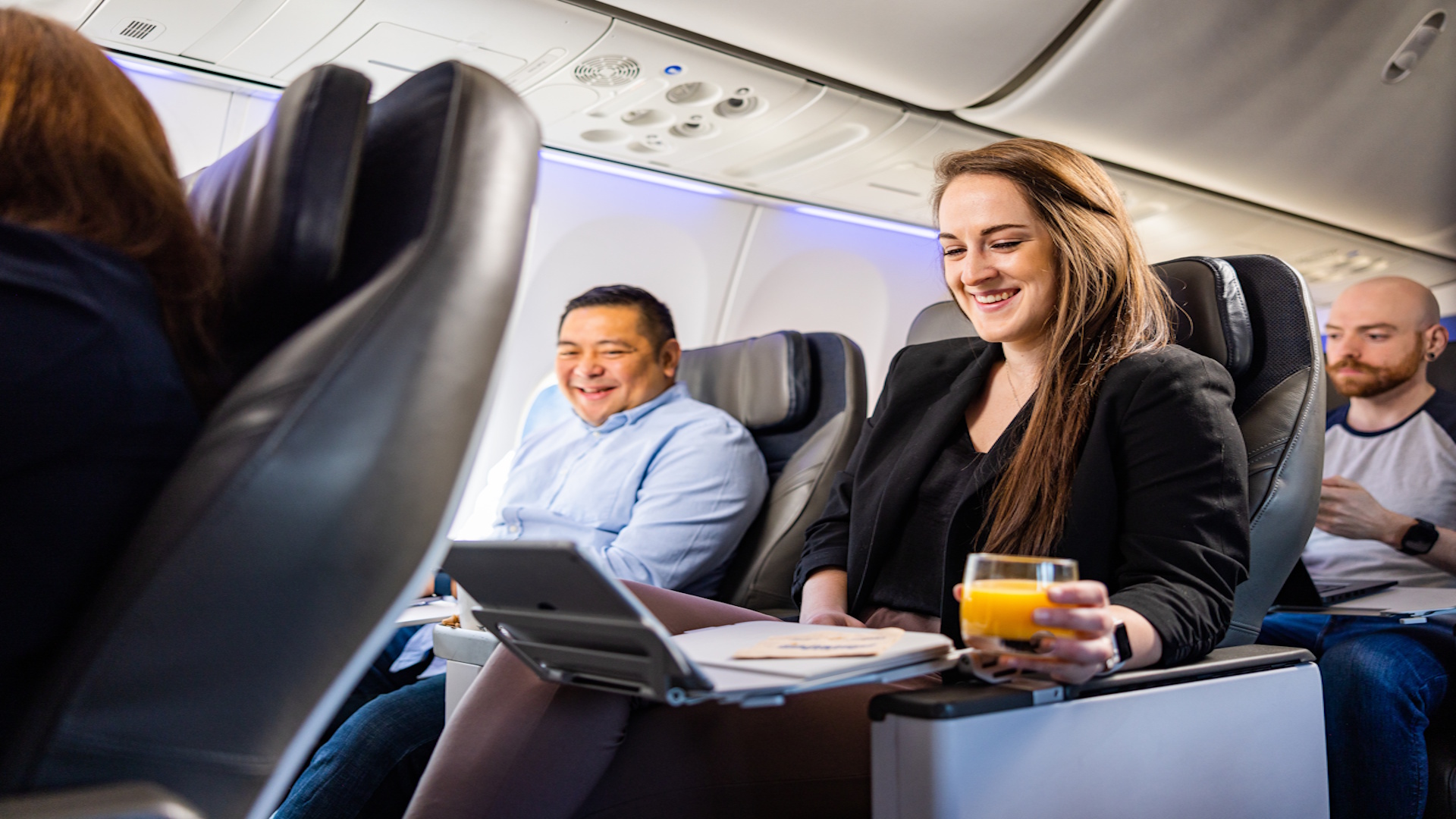 A woman using a laptop in Alaska Airlines Premium First Class, with a man looking at her screen and smiling from the window seat.