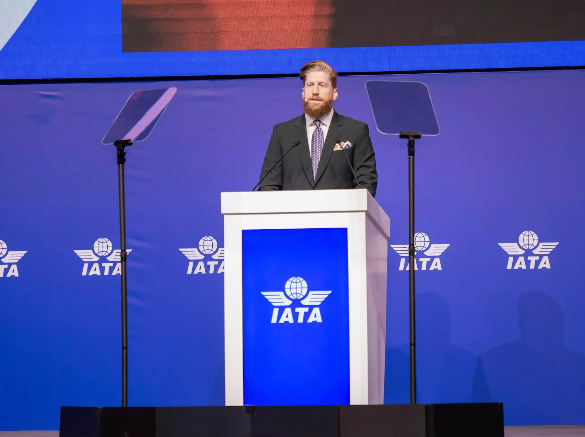 An image of a bearded man standing behind a podium at an International Air Transport Association (IATA) conference.