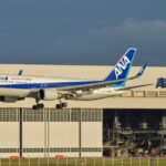 An All Nippon Airways (ANA) Boeing 767-300ER airplane with landing gear down, flying low in front of a large white ANA maintenance hangar. The aircraft features the "Inspiration of Japan" livery, and the registration JA622A is visible on the rear fuselage. A second, smaller ANA aircraft is parked inside the hangar in the background under a clear, late-afternoon sky.