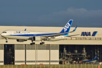 An All Nippon Airways (ANA) Boeing 767-300ER airplane with landing gear down, flying low in front of a large white ANA maintenance hangar. The aircraft features the "Inspiration of Japan" livery, and the registration JA622A is visible on the rear fuselage. A second, smaller ANA aircraft is parked inside the hangar in the background under a clear, late-afternoon sky.