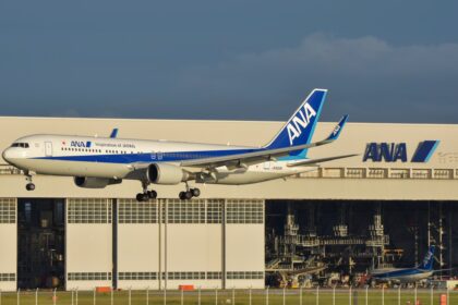 An All Nippon Airways (ANA) Boeing 767-300ER airplane with landing gear down, flying low in front of a large white ANA maintenance hangar. The aircraft features the "Inspiration of Japan" livery, and the registration JA622A is visible on the rear fuselage. A second, smaller ANA aircraft is parked inside the hangar in the background under a clear, late-afternoon sky.