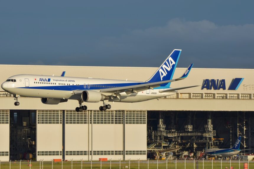An All Nippon Airways (ANA) Boeing 767-300ER airplane with landing gear down, flying low in front of a large white ANA maintenance hangar. The aircraft features the "Inspiration of Japan" livery, and the registration JA622A is visible on the rear fuselage. A second, smaller ANA aircraft is parked inside the hangar in the background under a clear, late-afternoon sky.