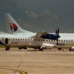 A side-view of a MASwings ATR 72 turboprop aircraft parked on a sunlit tarmac under a moody, dark sky. The plane features a white fuselage with a vibrant green and blue "hibiscus" design on the tail and the registration 9M-MWE. In the background, the tail of a larger Malaysia Airlines jet is visible, along with ground service vehicles and cargo carts.