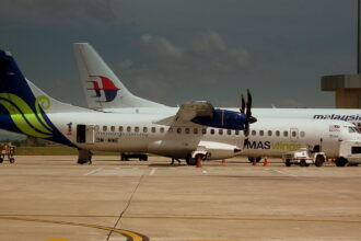 A side-view of a MASwings ATR 72 turboprop aircraft parked on a sunlit tarmac under a moody, dark sky. The plane features a white fuselage with a vibrant green and blue "hibiscus" design on the tail and the registration 9M-MWE. In the background, the tail of a larger Malaysia Airlines jet is visible, along with ground service vehicles and cargo carts.
