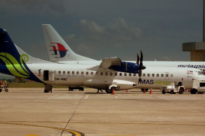 A side-view of a MASwings ATR 72 turboprop aircraft parked on a sunlit tarmac under a moody, dark sky. The plane features a white fuselage with a vibrant green and blue "hibiscus" design on the tail and the registration 9M-MWE. In the background, the tail of a larger Malaysia Airlines jet is visible, along with ground service vehicles and cargo carts.
