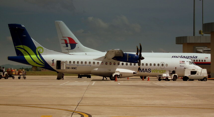 A side-view of a MASwings ATR 72 turboprop aircraft parked on a sunlit tarmac under a moody, dark sky. The plane features a white fuselage with a vibrant green and blue "hibiscus" design on the tail and the registration 9M-MWE. In the background, the tail of a larger Malaysia Airlines jet is visible, along with ground service vehicles and cargo carts.