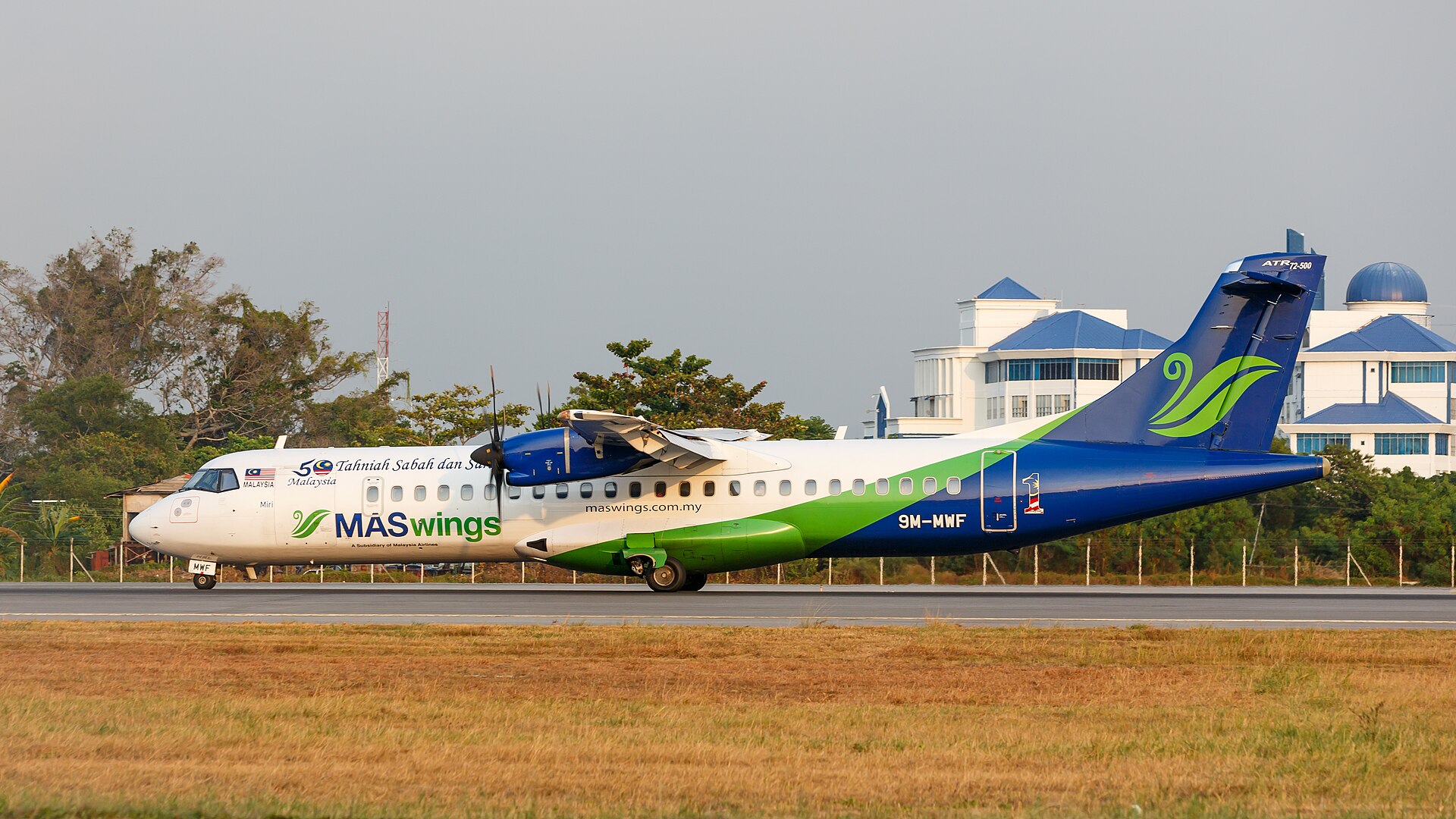 A full profile view of a MASwings ATR 72-500 turboprop aircraft, registration 9M-MWF, taxiing on a runway. The plane features a white fuselage transitioning into a bright green and blue wave pattern toward the tail. Special