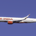 A side-profile view of an Air India Airbus A350 flying through a clear, solid blue sky. The aircraft features the airline's updated livery, with a white fuselage, bold red "AIR INDIA" lettering, and a vibrant tail fin decorated in shades of purple, red, and gold with a geometric pattern. The engines are bright red, and the curved wingtips (winglets) also feature the purple and gold design. Air India is currently undergoing a leadership reset, according to sources.