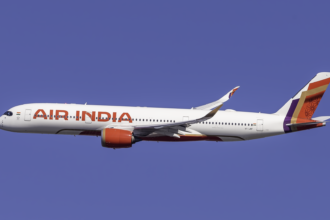 A side-profile view of an Air India Airbus A350 flying through a clear, solid blue sky. The aircraft features the airline's updated livery, with a white fuselage, bold red "AIR INDIA" lettering, and a vibrant tail fin decorated in shades of purple, red, and gold with a geometric pattern. The engines are bright red, and the curved wingtips (winglets) also feature the purple and gold design. Air India is currently undergoing a leadership reset, according to sources.