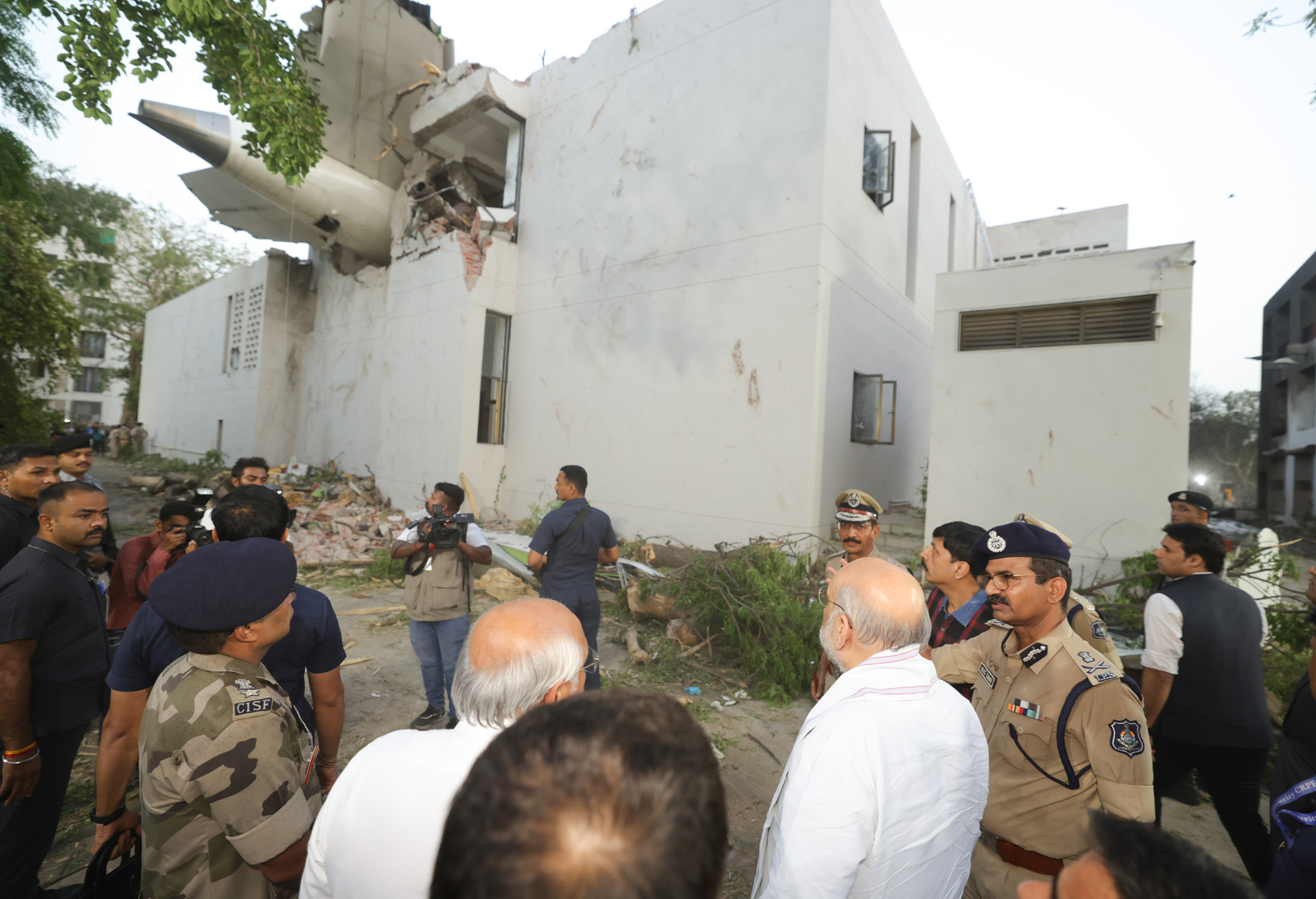 A wide shot showing the tail section of a large aircraft lodged into the corner of a white, two-story building. The impact has shattered the concrete and brickwork, with visible debris and tree branches on the ground. In the foreground, several men in police and military uniforms, some wearing CISF patches, are gathered and looking toward the damaged structure. A few civilians and a news cameraman are also present at the scene.