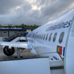 Side view of an Azores Airlines passenger jet parked on the apron at Ponta Delgada Airport in the Azores. The image shows the white fuselage of an Airbus A321neo, with a row of oval cabin windows running along the aircraft’s side and the airline name “azores airlines” printed in dark lettering. An open aircraft door with safety instructions is visible in the foreground, along with a Portuguese flag near the rear of the door area. The wing and one engine extend to the left, connected to a covered boarding walkway. In the background, the airport terminal building sits under a dramatic, cloud-filled sky, with ground crew vehicles faintly visible on the tarmac.