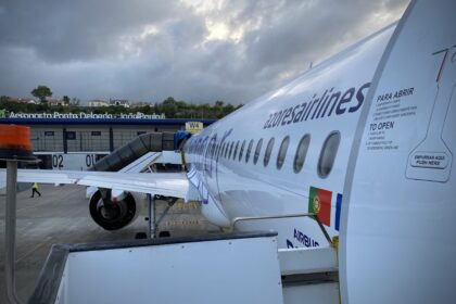 Side view of an Azores Airlines passenger jet parked on the apron at Ponta Delgada Airport in the Azores. The image shows the white fuselage of an Airbus A321neo, with a row of oval cabin windows running along the aircraft’s side and the airline name “azores airlines” printed in dark lettering. An open aircraft door with safety instructions is visible in the foreground, along with a Portuguese flag near the rear of the door area. The wing and one engine extend to the left, connected to a covered boarding walkway. In the background, the airport terminal building sits under a dramatic, cloud-filled sky, with ground crew vehicles faintly visible on the tarmac.