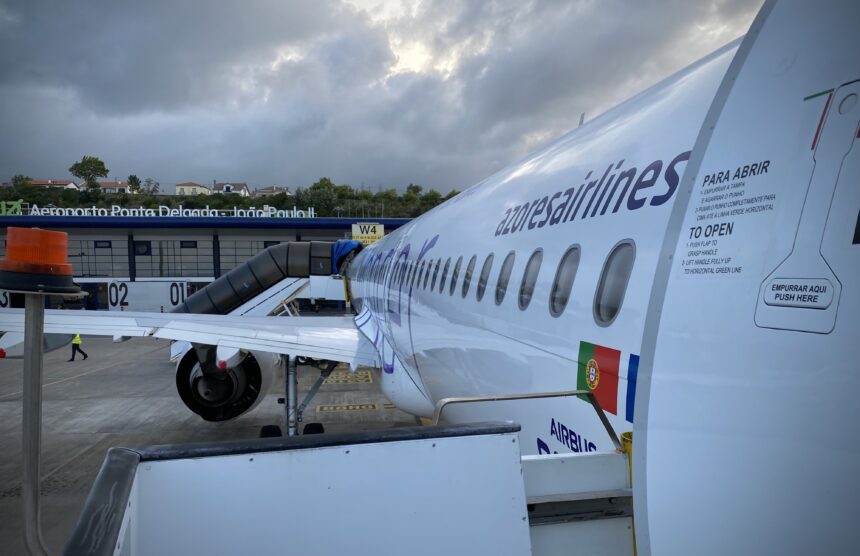 Side view of an Azores Airlines passenger jet parked on the apron at Ponta Delgada Airport in the Azores. The image shows the white fuselage of an Airbus A321neo, with a row of oval cabin windows running along the aircraft’s side and the airline name “azores airlines” printed in dark lettering. An open aircraft door with safety instructions is visible in the foreground, along with a Portuguese flag near the rear of the door area. The wing and one engine extend to the left, connected to a covered boarding walkway. In the background, the airport terminal building sits under a dramatic, cloud-filled sky, with ground crew vehicles faintly visible on the tarmac.