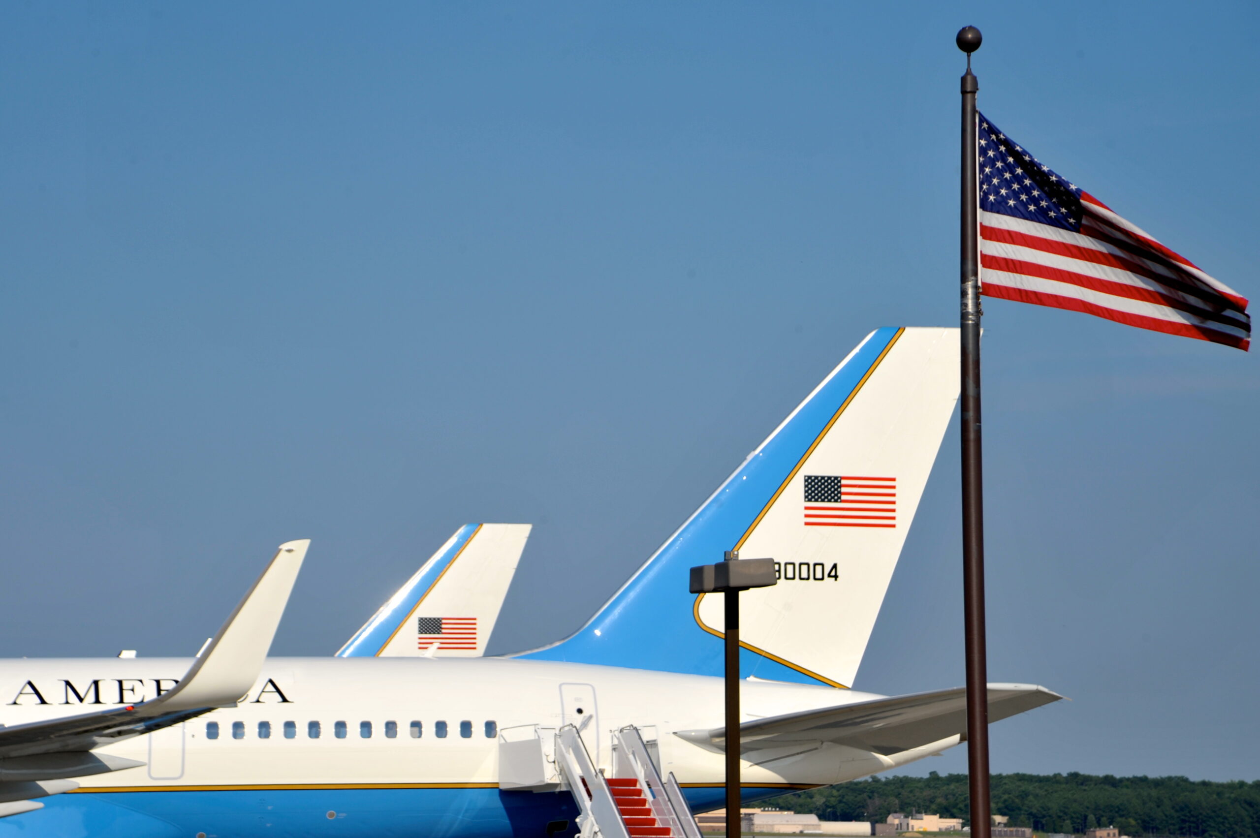 United States flag flying on a flagpole beside aircraft parked on an airfield, with aircraft tail fins visible in the background.