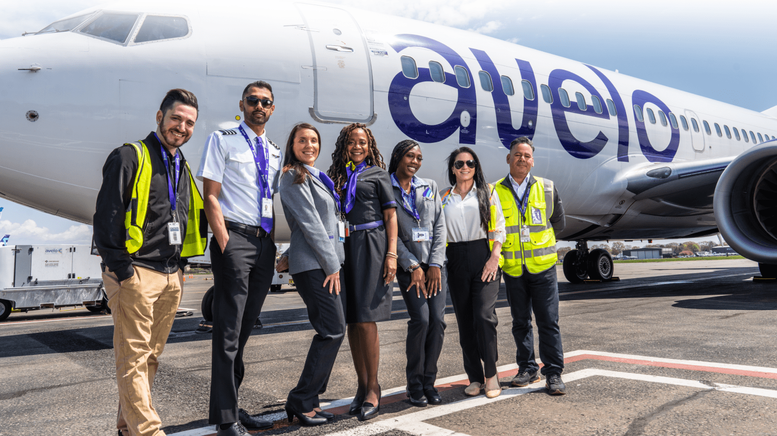 Members of Avelo Airlines crew stand on the tarmac in front of a parked Avelo aircraft and clear sky.