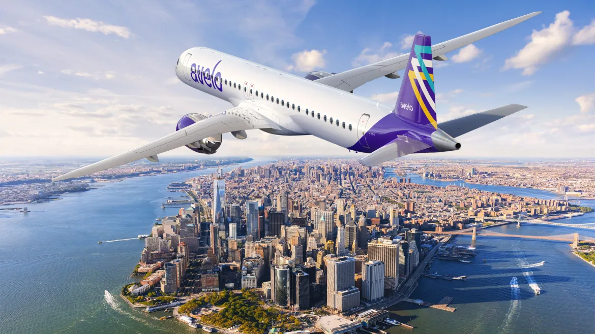 An Avelo Airlines aircraft flies over New York, with blue skies, dense buildings, surrounding greenery, and winding rivers visible below.