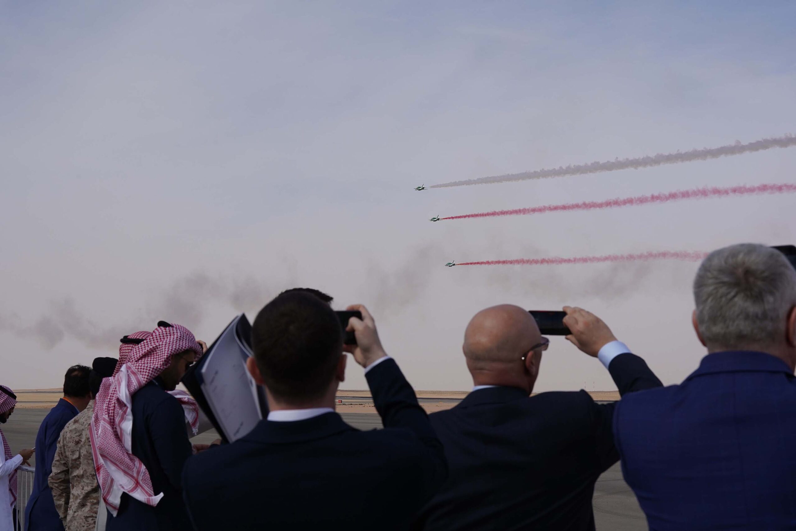 A view from behind a crowd of spectators at the World Defense Show in Saudi Arabia. Against a bright, clear sky, three green military jets fly in tight formation, leaving vibrant red smoke trails behind them. In the foreground, men in both professional business attire and traditional Saudi clothing are seen holding up their phones to record the flight display. The setting is a wide, open airfield at a purpose-built defence exhibition centre near Riyadh.