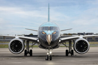 A front-on view of an Embraer E195-E2 commercial aircraft parked on a tarmac. The nose of the plane features a highly detailed "Tech Eagle" livery, with stylised eagle eyes and a beak painted over the cockpit and nose cone.